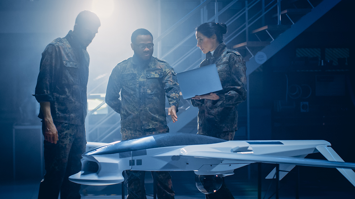 Army Aerospace Engineers Work On Unmanned Aerial Vehicle / Drone. Uniformed Aviation Experts Talk, Using Laptop. Industrial Facility with Aircraft for: Surveillance, Warfare Tactics, Air Strike
